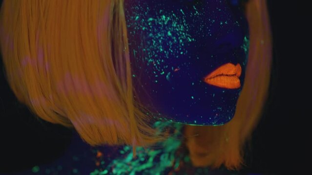 Creative body art. Close up portrait of young lady with glowing space make up wearing neon orange wig, tracking shot