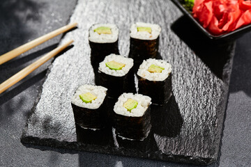 Avocado hosomaki on dark stone table. Hosomaki with avocado. Sushi roll with avocado inside. Style concept japanese menu with black background, leaves and hard shadow.