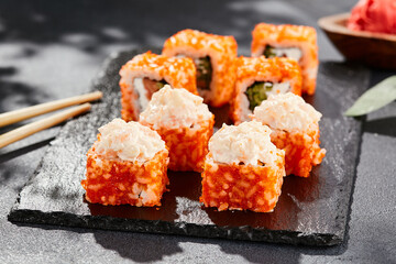 Maki sushi on dark stone table. California maki with tobiko. Sushi roll with salmon, cucumber inside, maguro, crab outside. Style concept japanese menu with black background, leaves and hard shadow.