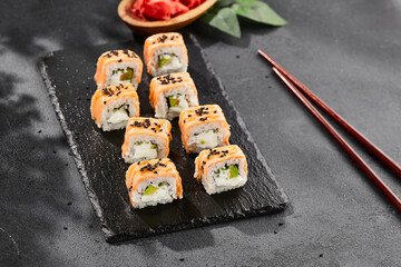 Maki sushi on dark slate. Hot philadelphia maki with baked salmon. Sushi roll with grilled salmon, cream cheese and avocado. Style concept japanese menu with black background, leaves and hard shadow.
