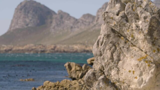 A beautiful uninhabited seashore rocky outcrop with rack focus to mountain peaks and blue sea in the background. Daytime.