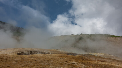 The soil in the geothermal valley is covered with orange sulfur deposits. Steam and smoke from fumaroles rise into the blue sky. The slopes of the mountains are hidden in a haze. Kamchatka
