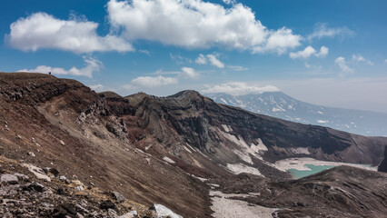 Acid lake on top of a volcano. Snow melting on steep slopes. The layered structure of the soil. On the edge of the crater, against the sky and clouds, tiny silhouettes of people are visible. Kamchatka