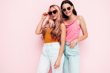 Two young beautiful smiling brunette hipster female in trendy summer clothes. carefree women posing near pink wall. Positive models having fun. Cheerful and happy. In sunglasses