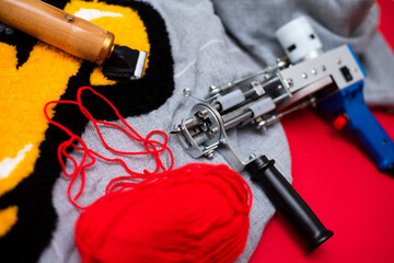 The tufting gun lies on a gray burlap, ready to work with the red yarn in the needle and the...