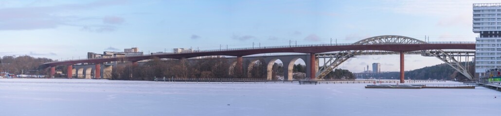 Panorama view of two train bridges, old and new, between the districts Årstadal and Södermalm, a snow covered and frozen lake Årstaviken a cold sunny winter day in Stockholm