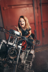 Fototapeta na wymiar Stylish baby brunette in a leather dress showing her tongue against the background of the garage. bikers free ride