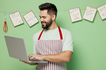 Young happy fun smiling male chef confectioner baker man 20s in striped apron hold use work on laptop pc computer isolated on plain pastel light green background studio portrait Cooking food concept