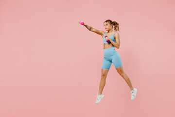 Fototapeta na wymiar Full size young strong sporty fitness trainer instructor woman wear blue tracksuit spend time in home gym jump high hold dumbbells isolated on pastel plain light pink background Workout sport concept