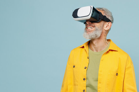 Elderly fun amazed happy gray-haired mustache bearded man 50s in yellow shirt watching in vr headset pc gadget isolated on plain pastel light blue background studio portrait. People lifestyle concept.
