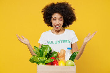 Young woman of African American ethnicity wears white volunteer t-shirt hold box with food vegetables spread hands isolated on plain yellow background. Voluntary free work help charity grace concept