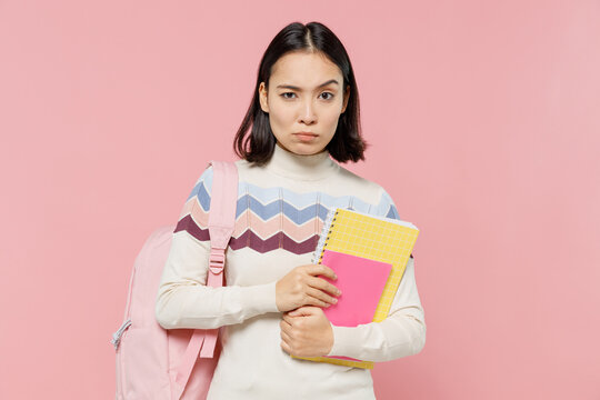 Serious strict sad teen student girl of Asian ethnicity wearing sweater backpack hold books look camera isolated on pastel plain light pink color background Education in university college concept