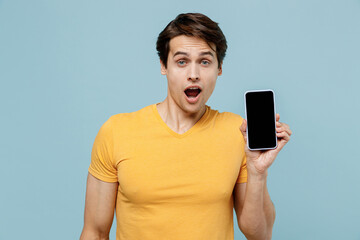 Young shocked amazed man 20s wearing yellow t-shirt holding in hand use mobile cell phone with blank screen workspace area copy space isolated on plain pastel light blue background studio portrait