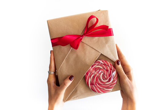 Beautifully wrapped gift in kraft paper with a red bow and a bright sweet candy for decoration in the hands of a girl. The girl holds a gift in her hands, photo on a white clean background, top view