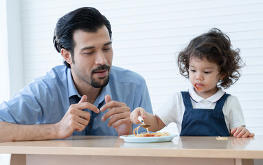 Caucasian young father with beard looking and cheer up little cute daughter trying to eat spaghetti with spoon by herself at home and adorable kid girl enjoy eating with face is mess up with ketchup