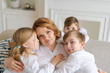 Obraz na płótnie Canvas We love you. Smiling young woman, kids hug mom while sitting on sofa in living room at home. Happy caucasian family enjoying a day together in a bright room