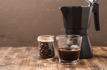 Glass of cofee with cofee beans and black  maker. Selective focus. Cup of black coffee in a glass and grains in a glass jar with a coffee maker on a brown background.