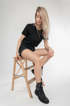 Sexy pretty blonde woman in fashion black clothes with t-shirt, shorts and leather shoes sits on a wooden chair. Beauty girl model in studio