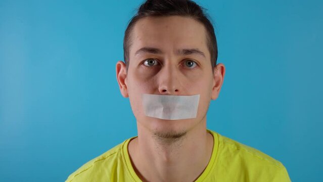 A young millennial guy is taping his mouth shut. Blue background. Ban on conversations