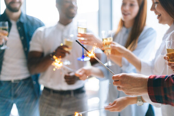 Close-up cropped shot of happy business team of young multi-ethnic startup businesspeople holding Bengal lights and clinking glasses of champagne at corporate party, on background of window.