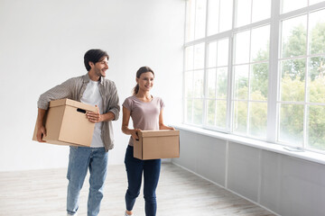 Fototapeta na wymiar Smiling young caucasian guy and lady carrying cardboard boxes in empty room of their own apartment