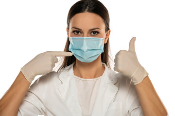 Beautiful female doctor or nurse wearing protective mask and latex or rubber gloves on white background with thumbs up