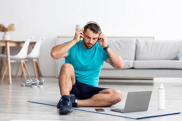 Serious millennial european male athlete puts headphones sits on mat in living room interior with bottle