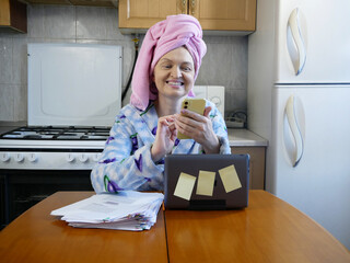 woman in bath towel on her head and wearing bathrobe working with laptop and documents in kitchen at home, woman accountant conducts an audit, home office