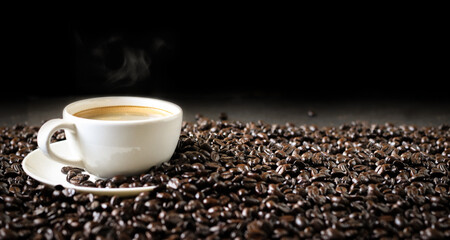 Hot coffee in a white coffee cup and many coffee beans placed around a on dark background.