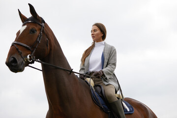 Fototapeta premium Beautiful young woman riding a horse outdoor. Concept of animal care. Rural rest and leisure. Idea of green tourism. Young european woman wearing helmet and uniform.