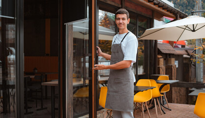 Fototapeta na wymiar barista or waiter cafe or coffee shop owner against entrance, gesture inviting you to visit, smiling guy in apron standing outdoors being proud of his small local business