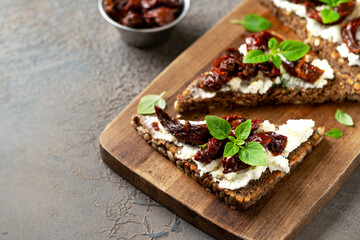 Bruschetta or whole grain bread sandwich with cream cheese and sun-dried tomatoes. Crostini on a wooden serving board on a dark kitchen table closeup	