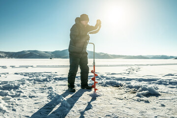 Fisherman drills a hole in the ice of a large frozen lake on a sunny day. The joy of winter fishing