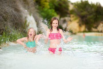 Two young sisters bathing in natural swimming pool in Bagno Vignoni, with thermal spring water and waterfall. Tuscany, Italy.