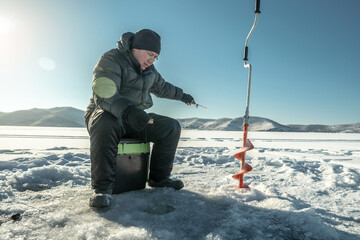 Fototapeta na wymiar Fisherman is fishing in a hole on the ice of a large frozen lake on a sunny day. The joy of winter fishing