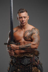 Muscular warrior with naked torso holding sword against gray background