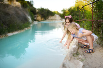 Two young sisters playing by natural swimming pool in Bagno Vignoni, with thermal spring water and waterfall. Tuscany, Italy.