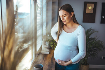 A young Caucasian pregnant woman with a headache is standing by the window in the living room. The...