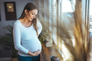 A young Caucasian pregnant woman with a headache is standing by the window in the living room. The concept of parenting expectations of pregnancy symptoms. 