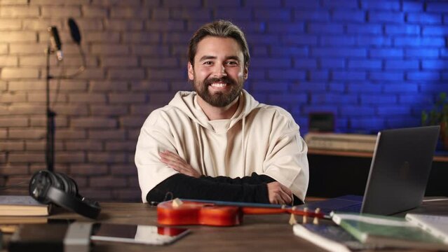 Portrait of handsome bearded man sitting at desk with modern laptop and ukulele, smiling and looking at camera. Male person spending free time for online studying in music sphere.