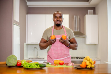 African man in an apron preparing food in the kitchen leaning two oranges against the rudy imitating female tits - obrazy, fototapety, plakaty