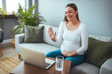 Pregnant woman video chat with family on laptop waving hand at screen. It shows how much the stomach has grown. Young pregnant woman with laptop browsing or chatting with friends online, sitting