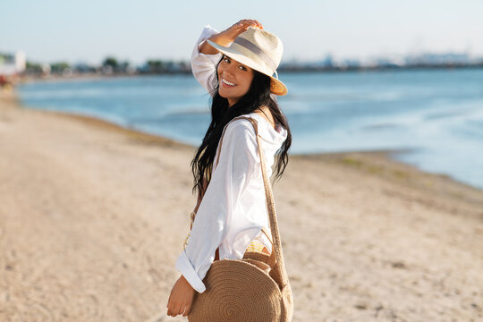 people, summer holidays and leisure concept - happy smiling woman in white shirt and straw hat with jute bag walking along beach