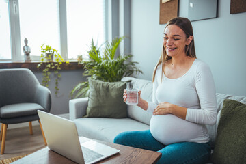 The pregnant woman is happy as she greets her family and friends on her laptop. She holds a glass of water while sitting on the sofa. The concept of a healthy lifestyle. 