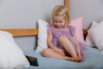  child sits on the bed watching a smartphone. A little blond girl in a lilac dress playing alone
