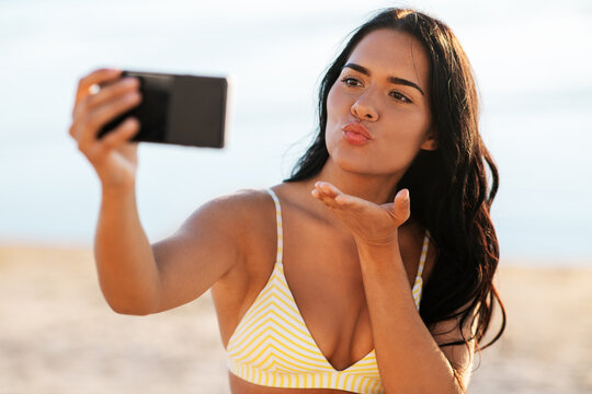 people, summer and swimwear concept - happy woman in bikini swimsuit taking selfie with smartphone and sending air kiss on beach