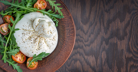 Top view of italian burrata white homemade soft milk cheese made of mozzarella and cream served with sliced cherry tomatoes and arugula leaves o plate on dark brown wooden background with copy space