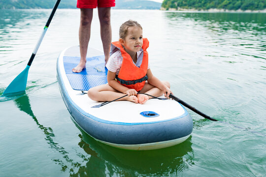 Close Up Of Cute Caucasian Girl In Swim Life Vest Swimming And Her Father On SUP Board. Family Paddleboarding On The Lake On Summer Day. Active Leisure With Kids. Family Local Getaway Concept