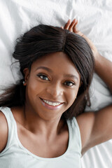 Glad happy pretty young african american lady lies on white comfortable bed and looks at camera in bedroom