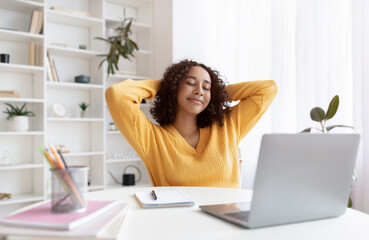 Relaxed young black female student sitting with hands behind head near laptop, having break after online class at home